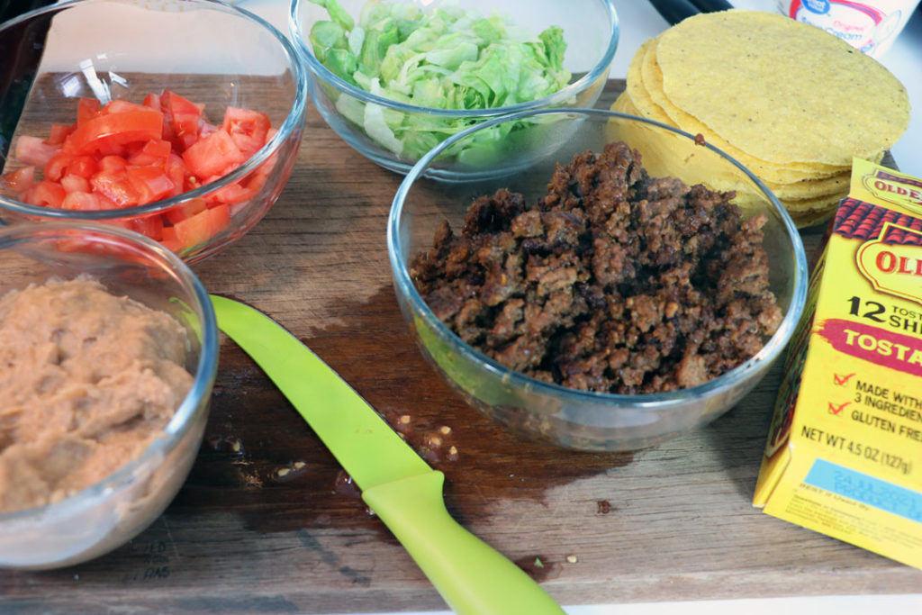 taco ingredients on a table. ground beef, refried beans, lettuce, shells, and tomatoes