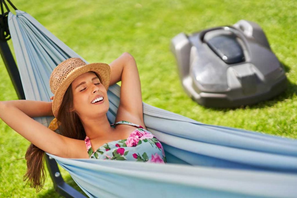 A homeowner relaxing on a patio with a robotic mower working in the background.