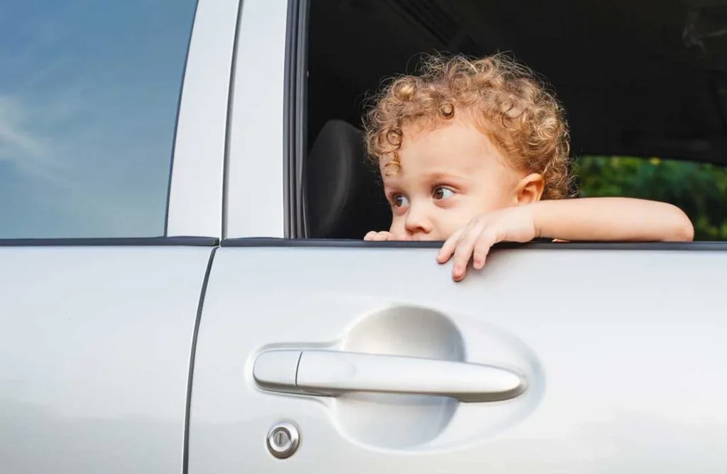A frustrated parent standing outside a locked car looking through the window, with young children visible inside or nearby, illustrating the common stress of a car lockout.