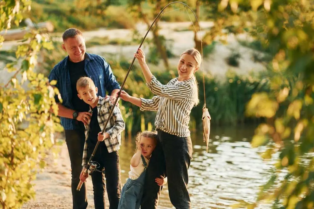 A happy family of four fishing together by a calm lake on a sunny day, emphasizing the joy of a family fishing trip.