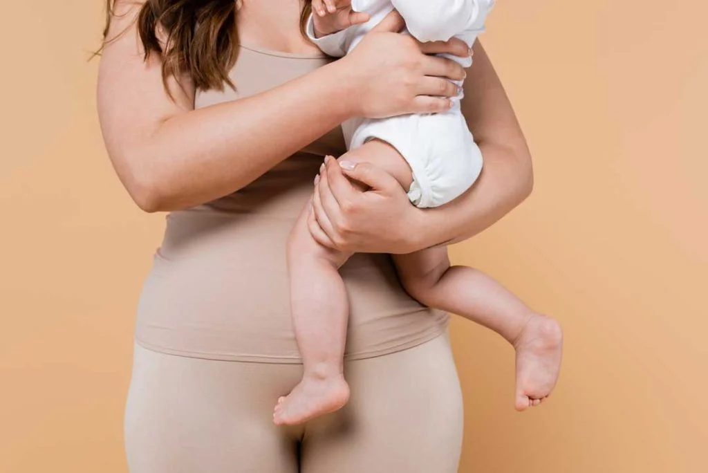 A new mother wearing a supportive abdominal compression garment while gently holding her baby, illustrating a tool for comfort and posture during C-section recovery.